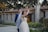 A couple is smiling and dancing together in an outdoor setting, surrounded by greenery and festive string lights. The bride wears a white dress and veil, and the groom is in a blue suit. The background features a stone building with a tiled roof.