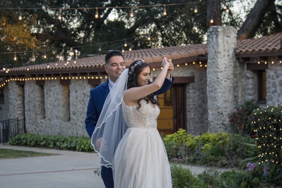 A couple is smiling and dancing together in an outdoor setting, surrounded by greenery and festive string lights. The bride wears a white dress and veil, and the groom is in a blue suit. The background features a stone building with a tiled roof.