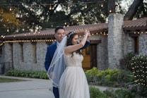 A couple is smiling and dancing together in an outdoor setting, surrounded by greenery and festive string lights. The bride wears a white dress and veil, and the groom is in a blue suit. The background features a stone building with a tiled roof.