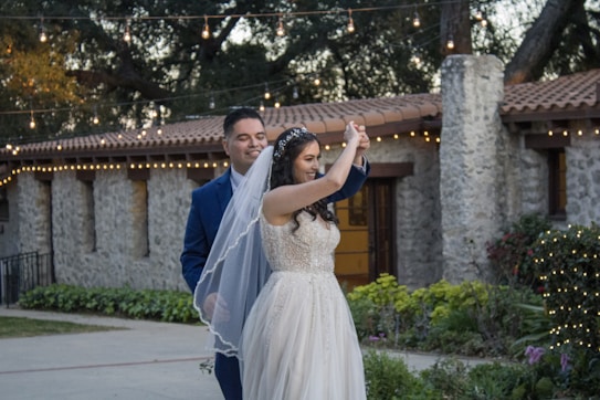 A couple is smiling and dancing together in an outdoor setting, surrounded by greenery and festive string lights. The bride wears a white dress and veil, and the groom is in a blue suit. The background features a stone building with a tiled roof.