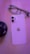 A sleek smartphone with a dual-camera setup is placed with its screen facing down, showcasing a metallic logo. The background is softly blurred, and a pair of eyeglasses and a round object are visible, all tinted with a purple hue.
