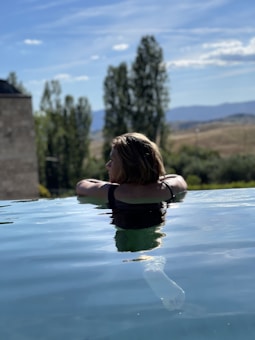 A person with shoulder-length hair is relaxing in an infinity pool, overlooking a scenic landscape with trees and distant hills under a clear blue sky.