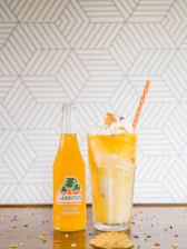 A glass bottle of mango-flavored Jarritos soda is placed next to a tall glass filled with an orange and cream float. The float is topped with whipped cream, colorful sprinkles, a small waffle cone, and a striped orange straw. The background features a geometric pattern, and colorful sprinkles are scattered on the wooden surface.