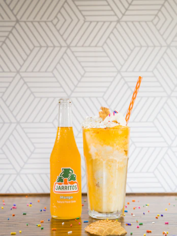 A glass bottle of mango-flavored Jarritos soda is placed next to a tall glass filled with an orange and cream float. The float is topped with whipped cream, colorful sprinkles, a small waffle cone, and a striped orange straw. The background features a geometric pattern, and colorful sprinkles are scattered on the wooden surface.