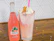 A glass filled with a creamy and colorful ice cream float, topped with whipped cream, sprinkles, and what appears to be a wafer. Next to it, there is a bottle of pink guava soda with the brand label visible. The background has a patterned white and gray wall, and the wooden surface is scattered with multicolored sprinkles.