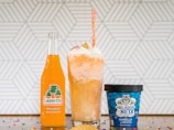 A vibrant orange Jarritos soda bottle, a tall float with whipped cream topped with colorful sprinkles, and a blue container of Mexican vanilla ice cream on a countertop decorated with scattered rainbow sprinkles. A geometric white and gray pattern serves as the background.