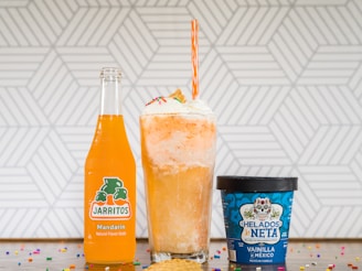 A vibrant orange Jarritos soda bottle, a tall float with whipped cream topped with colorful sprinkles, and a blue container of Mexican vanilla ice cream on a countertop decorated with scattered rainbow sprinkles. A geometric white and gray pattern serves as the background.