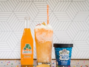 A vibrant orange Jarritos soda bottle, a tall float with whipped cream topped with colorful sprinkles, and a blue container of Mexican vanilla ice cream on a countertop decorated with scattered rainbow sprinkles. A geometric white and gray pattern serves as the background.