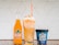A vibrant orange Jarritos soda bottle, a tall float with whipped cream topped with colorful sprinkles, and a blue container of Mexican vanilla ice cream on a countertop decorated with scattered rainbow sprinkles. A geometric white and gray pattern serves as the background.