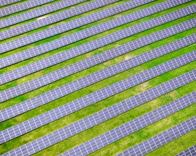 An array of solar panels arranged in parallel rows on a lush green field, capturing sunlight for energy conversion. The solar panels create a pattern of alternating blue and green stripes.