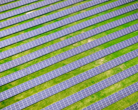 An array of solar panels arranged in parallel rows on a lush green field, capturing sunlight for energy conversion. The solar panels create a pattern of alternating blue and green stripes.