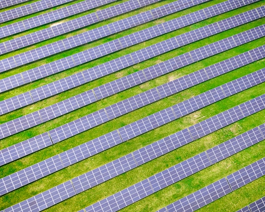 An array of solar panels arranged in parallel rows on a lush green field, capturing sunlight for energy conversion. The solar panels create a pattern of alternating blue and green stripes.