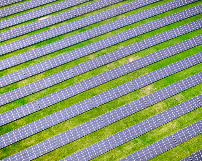 An array of solar panels arranged in parallel rows on a lush green field, capturing sunlight for energy conversion. The solar panels create a pattern of alternating blue and green stripes.