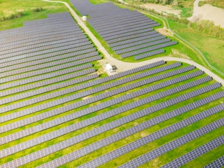 Rows of solar panels are arranged in a large open field, creating a pattern across the landscape. The panels are evenly spaced and set on a grassy terrain with some pathways cutting through the area.
