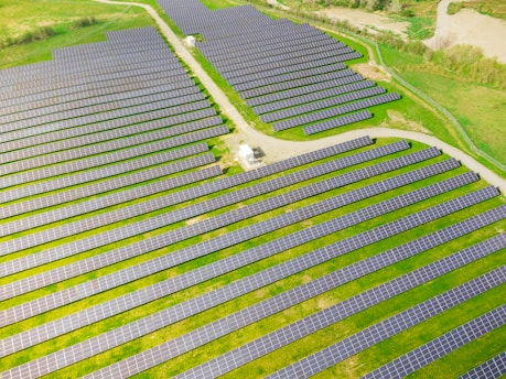 Rows of solar panels are arranged in a large open field, creating a pattern across the landscape. The panels are evenly spaced and set on a grassy terrain with some pathways cutting through the area.