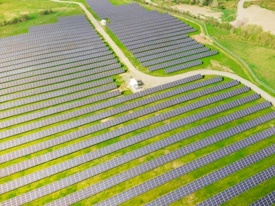 Rows of solar panels are arranged in a large open field, creating a pattern across the landscape. The panels are evenly spaced and set on a grassy terrain with some pathways cutting through the area.