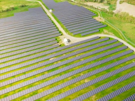 Rows of solar panels are arranged in a large open field, creating a pattern across the landscape. The panels are evenly spaced and set on a grassy terrain with some pathways cutting through the area.