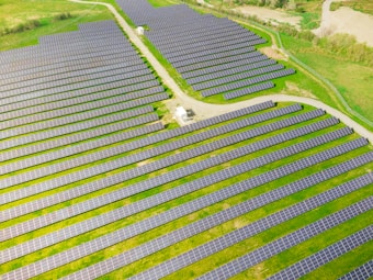 Rows of solar panels are arranged in a large open field, creating a pattern across the landscape. The panels are evenly spaced and set on a grassy terrain with some pathways cutting through the area.