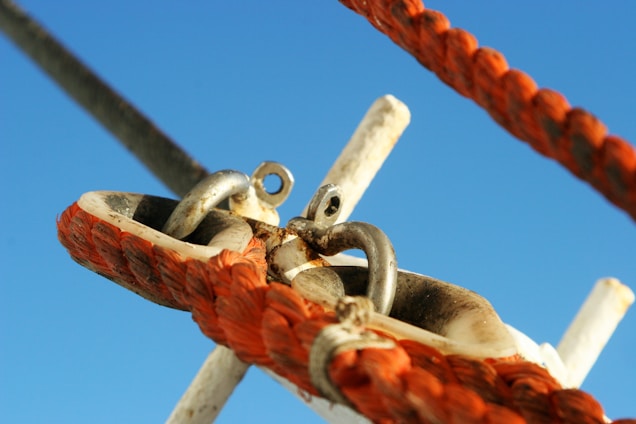 Portrait of a weathered and rusty metal shackle connected to an orange braided rope, captured against a clear blue sky. The ropes, bolts, and fastener hardware seem to be a part of a larger structure or mechanism, likely nautical in nature. There are signs of use and corrosion, suggesting the arrangement is exposed to harsh elements.