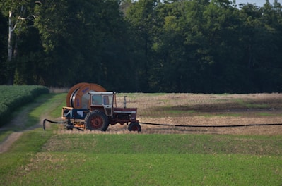 A red tractor with a large attached irrigation reel is positioned on a grassy path in a field landscape. The background consists of dense, dark green trees, while the foreground displays cultivated fields with patches of green and brown earth.