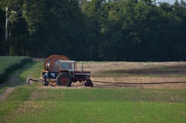 A red tractor with a large attached irrigation reel is positioned on a grassy path in a field landscape. The background consists of dense, dark green trees, while the foreground displays cultivated fields with patches of green and brown earth.