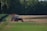A red tractor with a large attached irrigation reel is positioned on a grassy path in a field landscape. The background consists of dense, dark green trees, while the foreground displays cultivated fields with patches of green and brown earth.