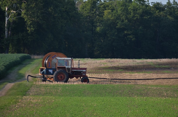 A  tractor with a large attached irrigation reel is positioned on a grassy path in a field landscape. The background consists of dense, dark green trees, while the foreground displays cultivated fields with patches of green and brown earth.