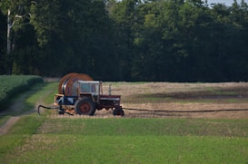 A red tractor with a large attached irrigation reel is positioned on a grassy path in a field landscape. The background consists of dense, dark green trees, while the foreground displays cultivated fields with patches of green and brown earth.