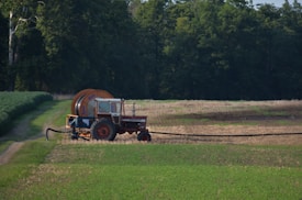 A red tractor with a large attached irrigation reel is positioned on a grassy path in a field landscape. The background consists of dense, dark green trees, while the foreground displays cultivated fields with patches of green and brown earth.