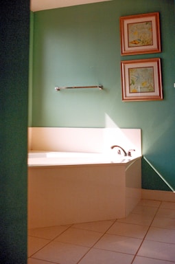 A bathroom corner with a large white bathtub featuring a chrome faucet. The walls are painted green and adorned with two framed artworks, one above the other. A chrome towel bar is also mounted on the wall. Sunlight casts a diagonal shadow across the bathtub and wall.