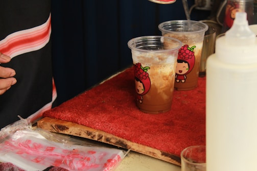Two plastic cups filled with a brown beverage, topped with a creamy layer, sit on a red textured mat. The cups feature a cute character design with a smiling face. A hand is partially visible on the left side, and another container with a spout is on the right.