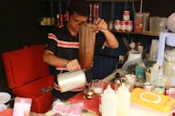 A person is preparing a drink using a cloth filter in a small kitchen setup. The workspace is cluttered with various bottles, jars, and utensils. The shelves behind are stocked with canned drinks and other supplies.