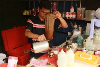 A person is preparing a drink using a cloth filter in a small kitchen setup. The workspace is cluttered with various bottles, jars, and utensils. The shelves behind are stocked with canned drinks and other supplies.