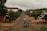A rural street scene featuring a dirt road lined with trees and modest buildings. A wooden cart loaded with blue bags is being pulled by a vehicle, with a few people present on the road and sidewalks. The sky is overcast, and the overall atmosphere appears calm and quiet.