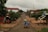 A rural street scene featuring a dirt road lined with trees and modest buildings. A wooden cart loaded with blue bags is being pulled by a vehicle, with a few people present on the road and sidewalks. The sky is overcast, and the overall atmosphere appears calm and quiet.