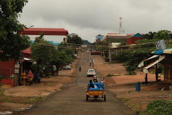 A rural street scene featuring a dirt road lined with trees and modest buildings. A wooden cart loaded with blue bags is being pulled by a vehicle, with a few people present on the road and sidewalks. The sky is overcast, and the overall atmosphere appears calm and quiet.