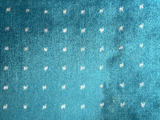 An image of a blue-green textured fabric with small, evenly spaced white dots scattered across the surface.