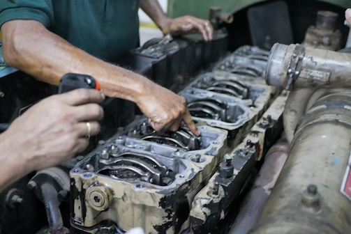 Hands are working on a disassembled engine with visible pistons and connecting rods. One hand is holding a flashlight while another is pointing at a part of the engine.