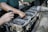 Hands are working on a disassembled engine with visible pistons and connecting rods. One hand is holding a flashlight while another is pointing at a part of the engine.