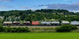 A freight train is moving along a railway track set against a lush, green landscape with dense forested hills. The train consists of various tank and cargo containers, some labeled with logos and text like 'Hamburg Sud'. Overhead, a partially cloudy sky adds depth to the scene.