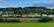 A freight train is moving along a railway track set against a lush, green landscape with dense forested hills. The train consists of various tank and cargo containers, some labeled with logos and text like 'Hamburg Sud'. Overhead, a partially cloudy sky adds depth to the scene.