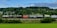 A freight train is moving along a railway track set against a lush, green landscape with dense forested hills. The train consists of various tank and cargo containers, some labeled with logos and text like 'Hamburg Sud'. Overhead, a partially cloudy sky adds depth to the scene.