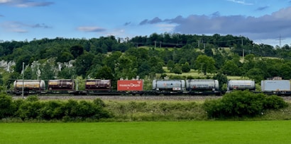 A freight train is moving along a railway track set against a lush, green landscape with dense forested hills. The train consists of various tank and cargo containers, some labeled with logos and text like 'Hamburg Sud'. Overhead, a partially cloudy sky adds depth to the scene.