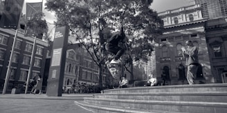 A skateboarder performs a trick in an urban setting in front of the State Library Victoria. There are trees and several people observing or walking by. Modern and classical architectural buildings are visible in the background, along with bicycle stands and flags.