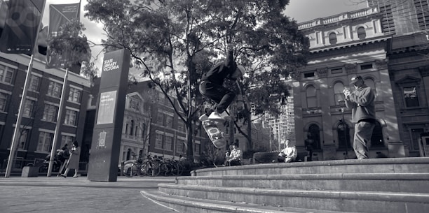 A skateboarder performs a trick in an urban setting in front of the State Library Victoria. There are trees and several people observing or walking by. Modern and classical architectural buildings are visible in the background, along with bicycle stands and flags.