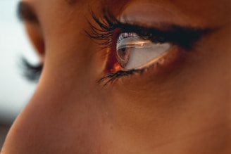 A close-up of a human eye with long eyelashes and soft lighting. The eye is well-defined, displaying detailed reflections of the surroundings. The surrounding skin appears smooth and slightly tanned.
