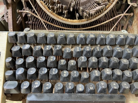 A vintage typewriter with a close-up view of its keyboard, featuring keys with unfamiliar characters. The machine shows signs of rust and wear, indicating its age and lack of use. The background reveals part of the inner mechanism with visible metal rods and circular components.