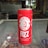 A red can labeled 'B Fizz' is placed on a glass surface in what appears to be a store setting. The text on the can mentions it is a fruit juice-based drink with malt flavor. The background shows a shelf, possibly with various products, and some natural light coming through a window.