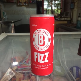 A red can labeled 'B Fizz' is placed on a glass surface in what appears to be a store setting. The text on the can mentions it is a fruit juice-based drink with malt flavor. The background shows a shelf, possibly with various products, and some natural light coming through a window.