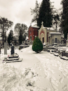A snow-covered cemetery with a variety of tombstones and a small chapel in the background. The chapel is made of red brick with white accents and has a cross on the roof. Surrounding the area are tall trees with no leaves, adding to the wintery atmosphere. There is a statue of a robed figure on a pedestal, next to a path partially covered with snow.